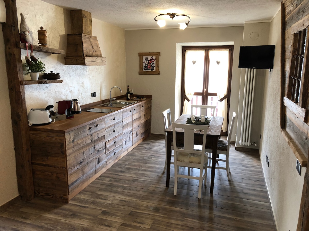 The kitchen area features rustic wooden cabinets and a countertop, complemented by modern appliances. A small dining table with four chairs is placed near a window that opens to a balcony, allowing natural light to illuminate the space.