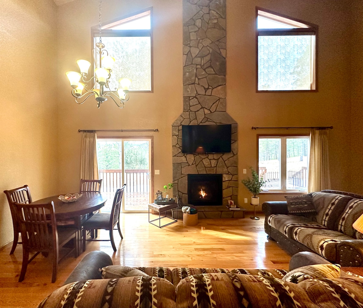 A spacious living area showcases a stone fireplace, framed by large windows that allow for natural light. A dining table accommodates multiple guests, while a comfortable couch invites relaxation. Outdoor views of the forest are visible through glass doors leading to the deck.
