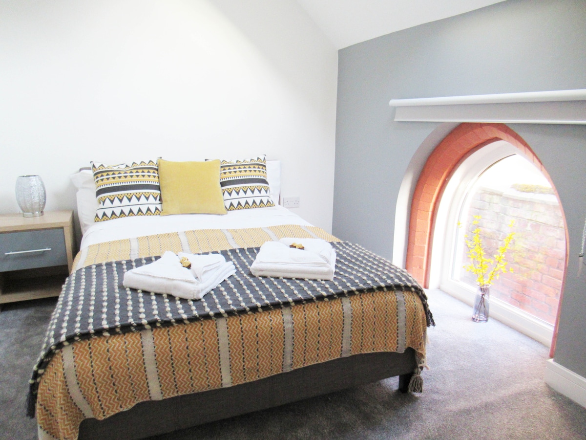 A comfortable bedroom features a spacious bed adorned with a patterned throw and neatly arranged towels. A yellow cushion adds a touch of colour. A unique arched window bathes the room in natural light, complemented by a small vase with yellow flowers.