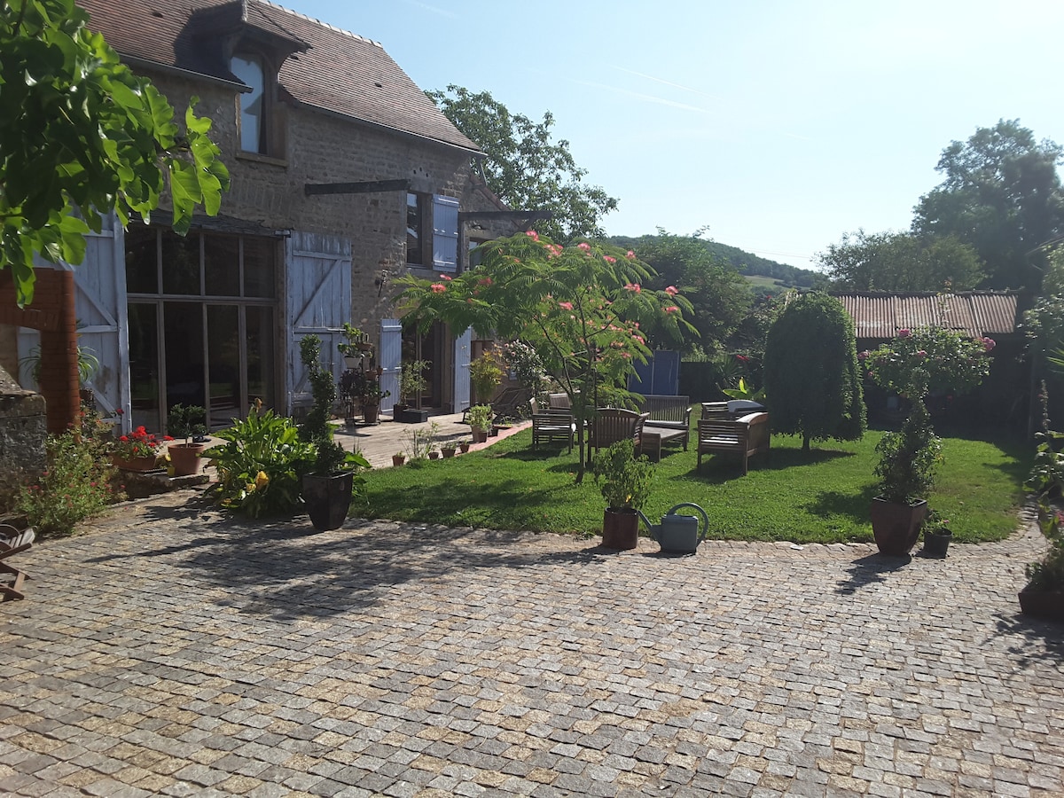 The image shows a sunlit outdoor area featuring a cobblestone path leading to a garden. Comfortable seating arrangements are present amidst lush greenery and various potted plants. The backdrop includes a well-maintained farmhouse with large glass doors, enhancing the serene countryside environment.