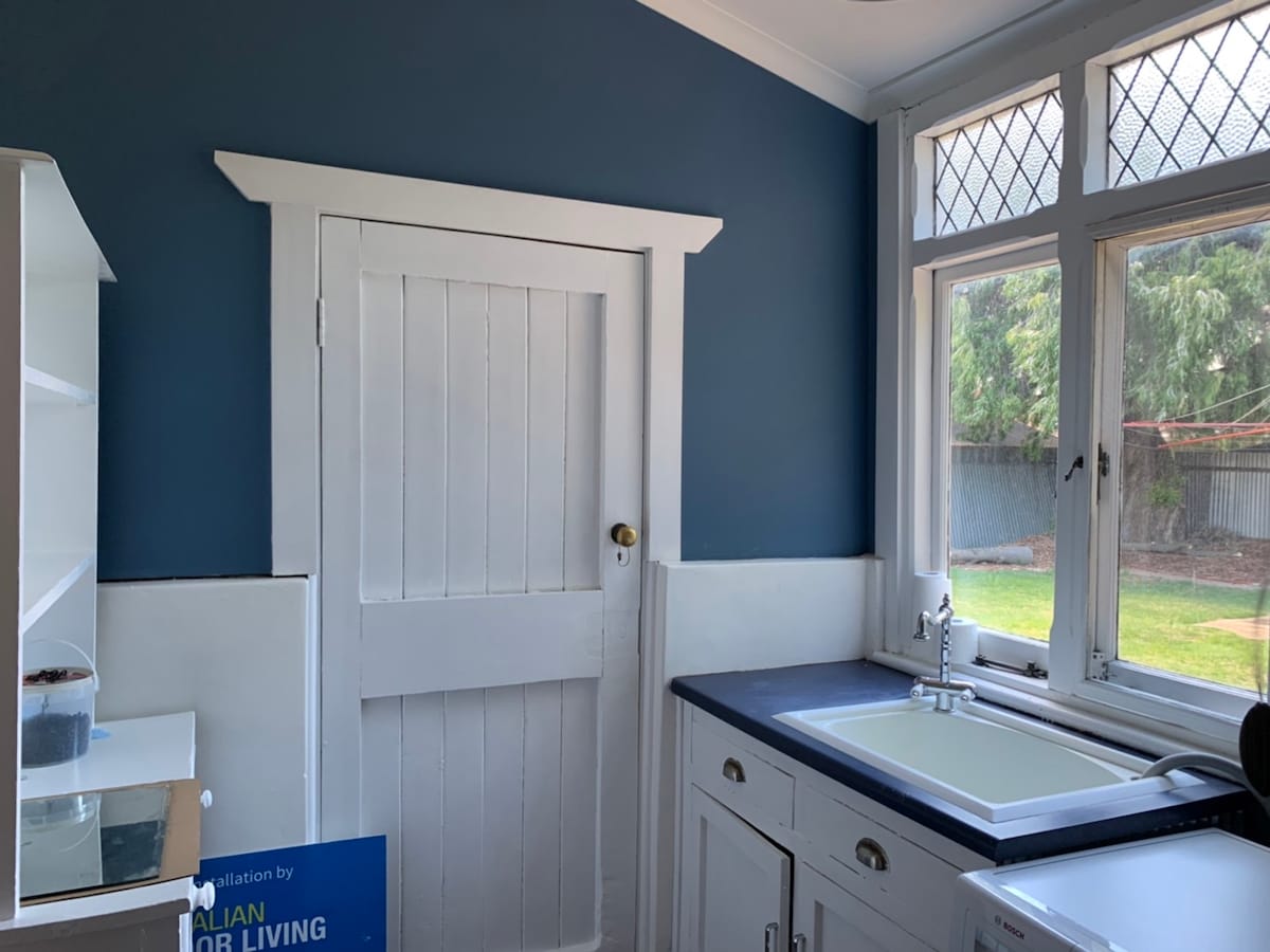 An interior view showcases a kitchen area with white cabinetry and a dark countertop. A door with a round brass doorknob leads to another room. Sunlight filters through the window, illuminating the space and highlighting the blue accent wall.