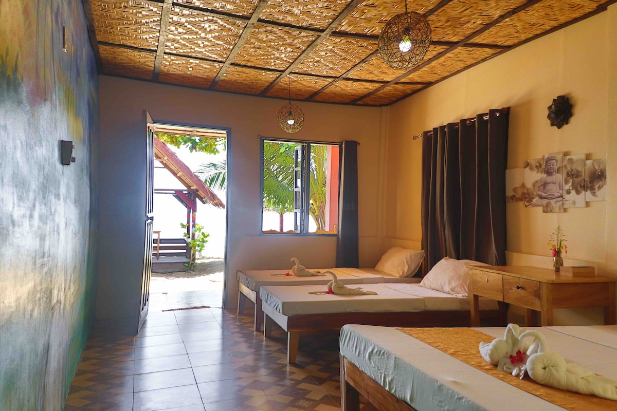 A bright room features three spacious beds arranged with decorative towel swans. Natural light enters through large windows, providing views of the outdoors. The ceiling is adorned with woven bamboo, and dark curtains frame the windows, offering a balance of light and privacy.