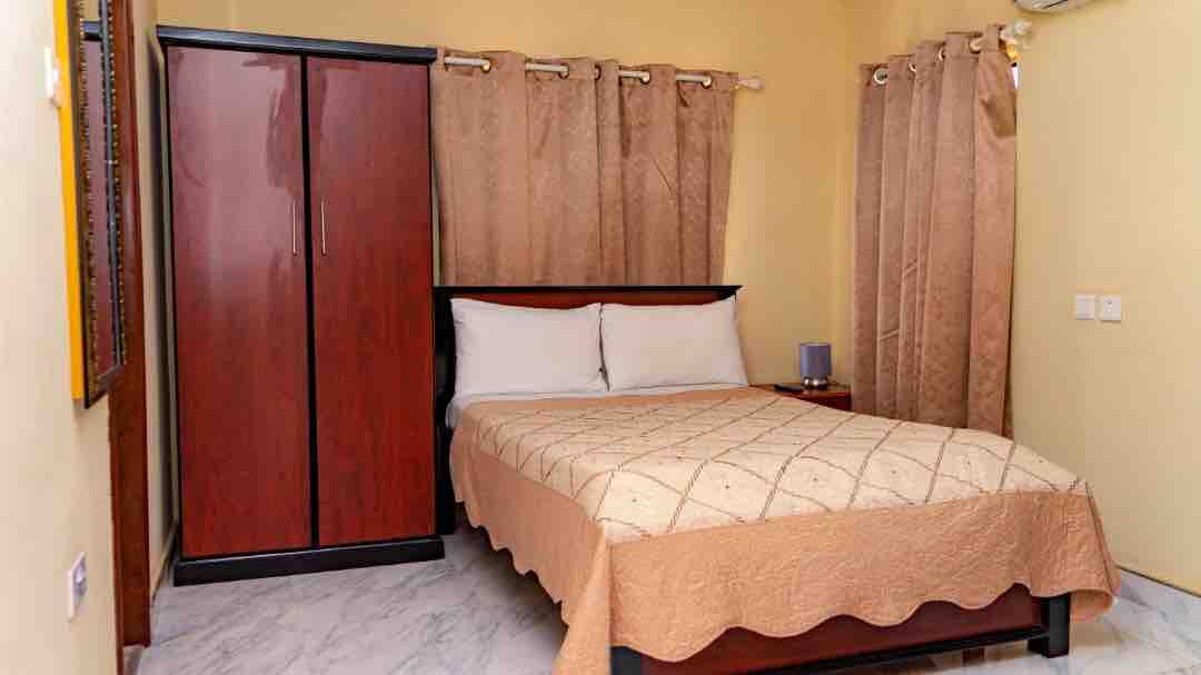 A comfortable sleeping area features a large bed with a light-colored quilt. A dark wood wardrobe stands adjacent to the bed, and soft curtains frame the window, allowing natural light to filter into the room. Neutral-colored walls create a calming atmosphere.