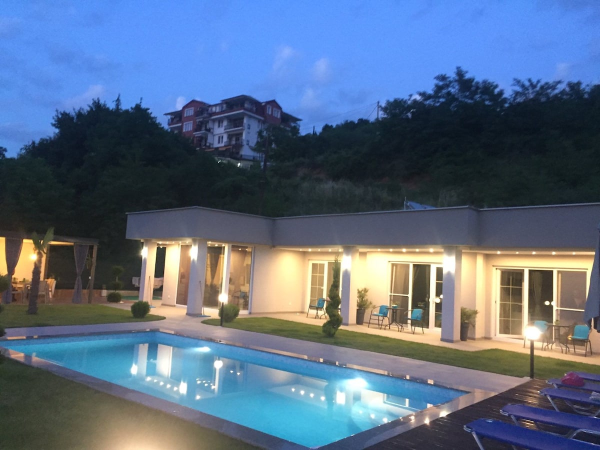 The villa exterior is illuminated in the evening, showcasing a clear swimming pool reflecting the sky. Comfortable seating areas are positioned around the pool, surrounded by greenery. The building features large glass doors, enhancing the connection to the outdoor space.