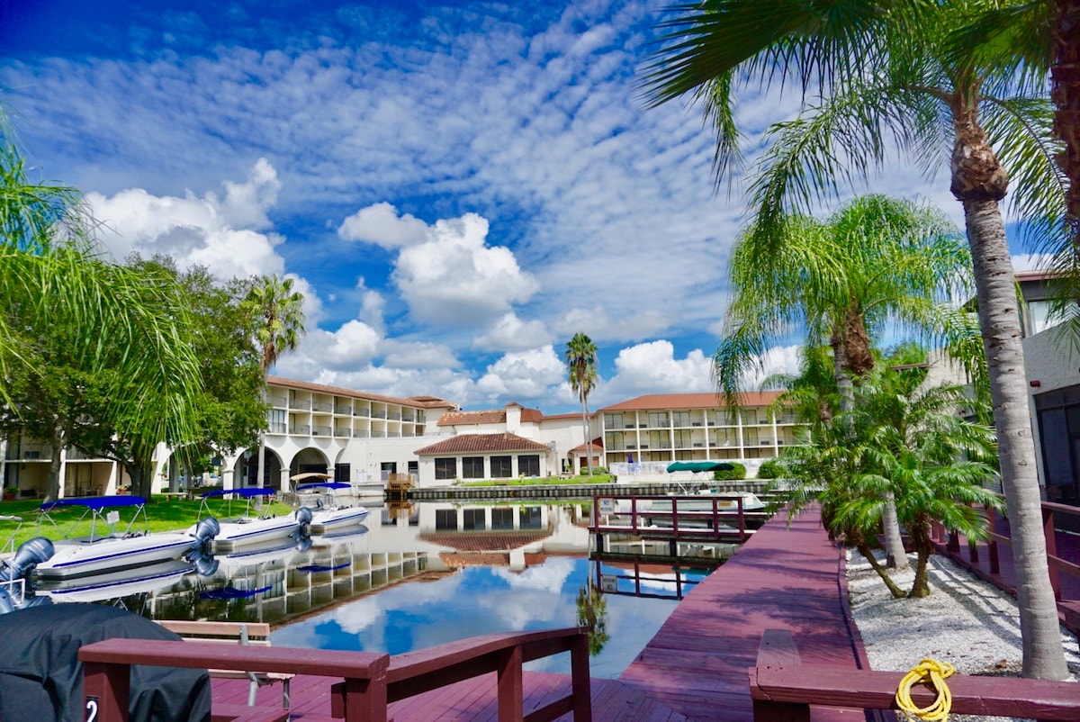 A serene waterfront view is presented, showcasing the calm lake reflecting the blue sky and fluffy clouds. The area is lined with vibrant palm trees and a wooden dock. The surrounding buildings feature a light-colored façade, creating a relaxed atmosphere.