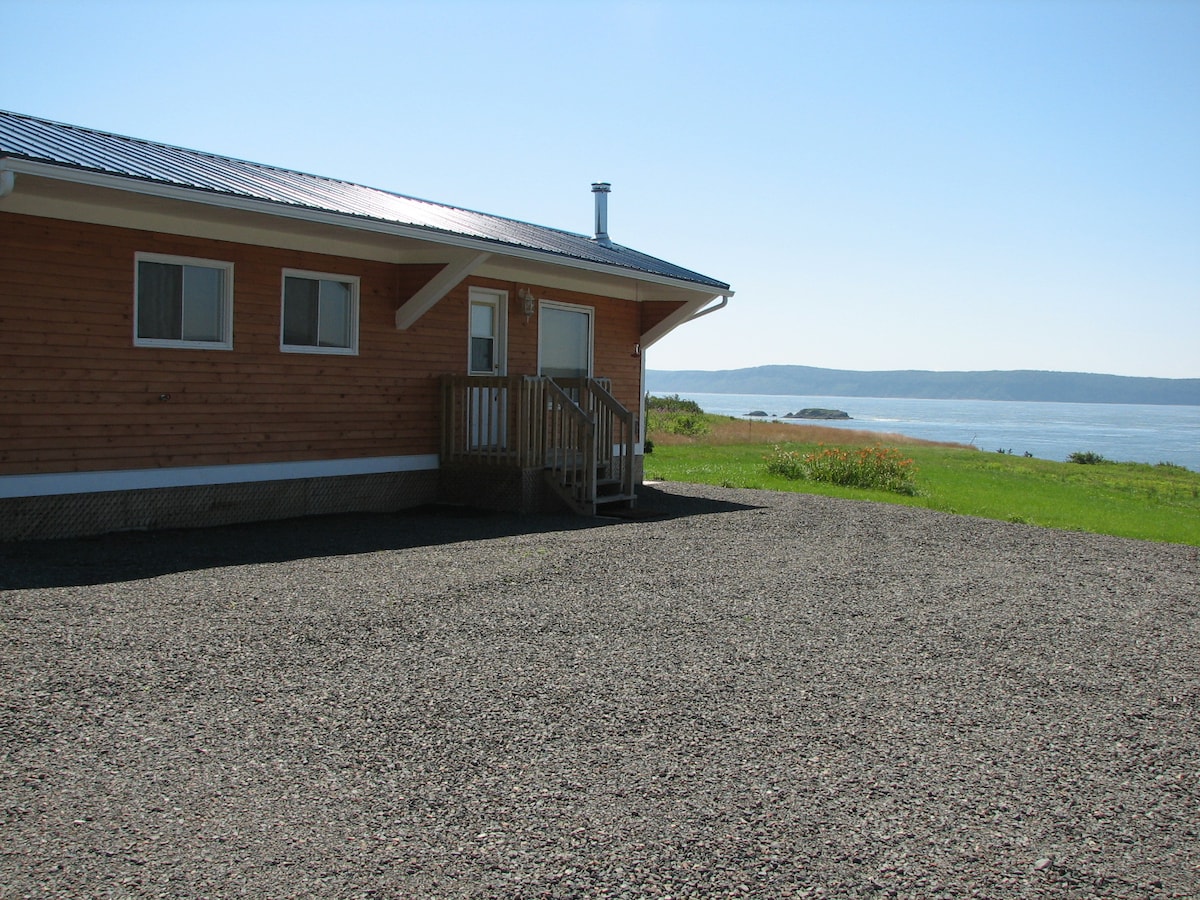 A wooden cottage is seen from the exterior, featuring a sloped roof and a small set of stairs leading to the entrance. The entrance is flanked by two windows. Ocean views are visible in the background, with a grassy area surrounding the cottage.