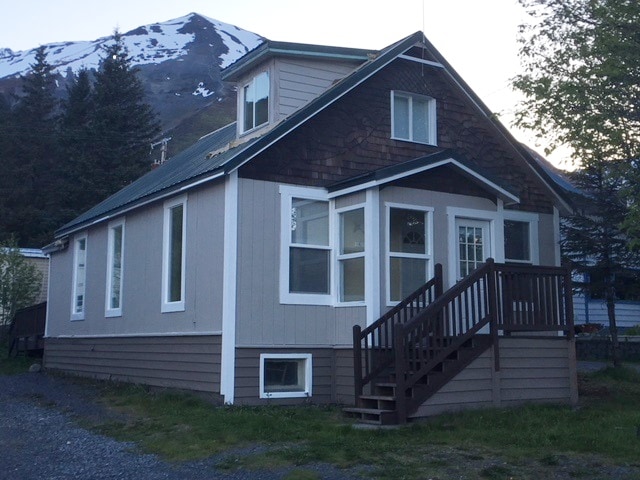 A quaint two-story cottage is visible with a peaked roof and wooden accents. Large windows allow light to filter in, while a welcoming front porch leads to a set of stairs. The backdrop features a mountainous landscape, enhancing the serene setting.