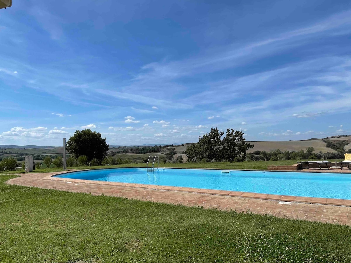 An outdoor swimming pool is surrounded by a well-maintained lawn, featuring a clear blue sky above. The pool area includes a tiled border and a distant view of rolling hills and greenery, creating a serene and spacious setting for relaxation.