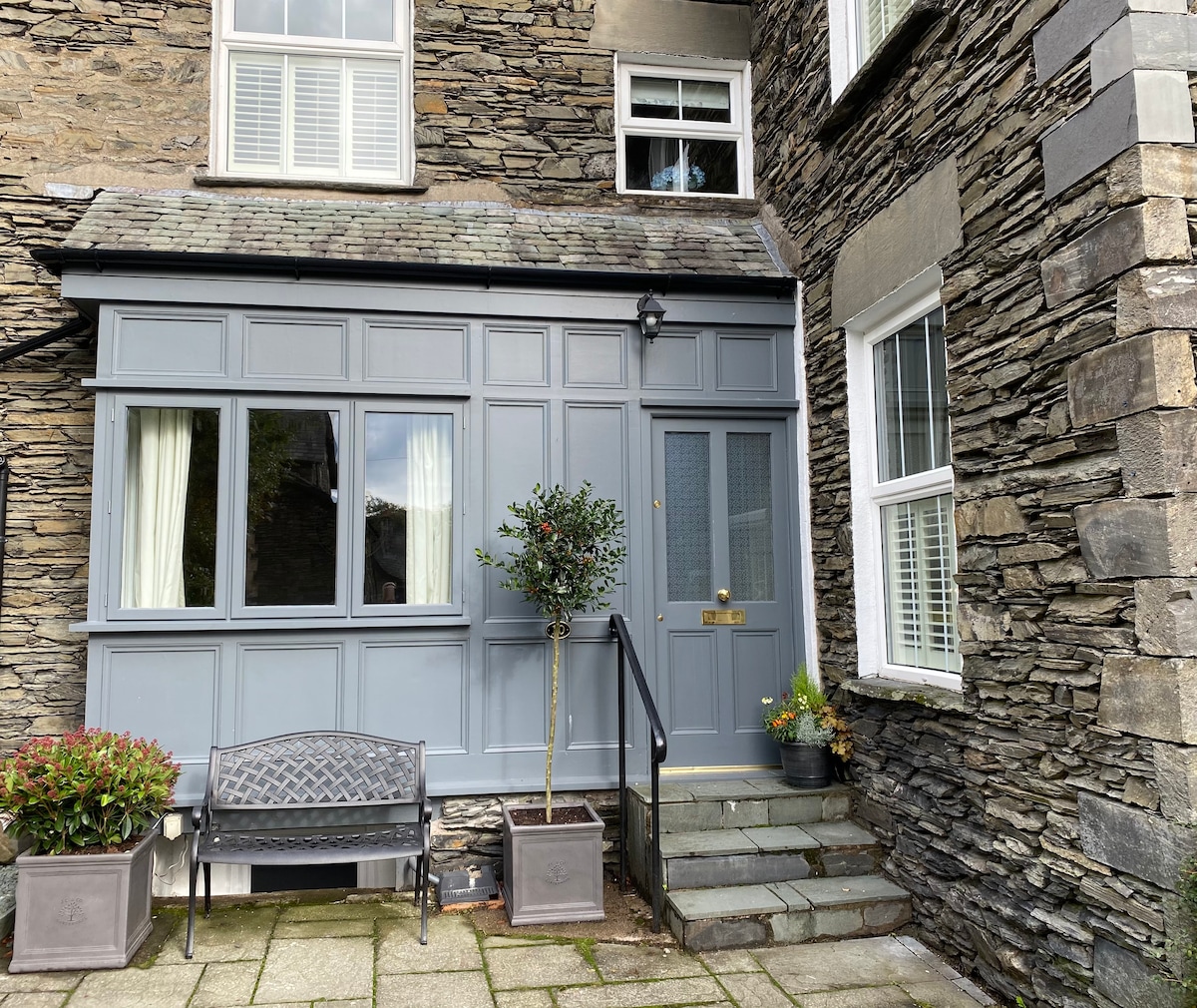 The entrance to the cottage features a slate stone facade with a grey panelled front door. Large windows framed in white allow natural light to enter. Two planters with greenery are placed alongside a low-profile bench and a set of stone steps leads to the door.