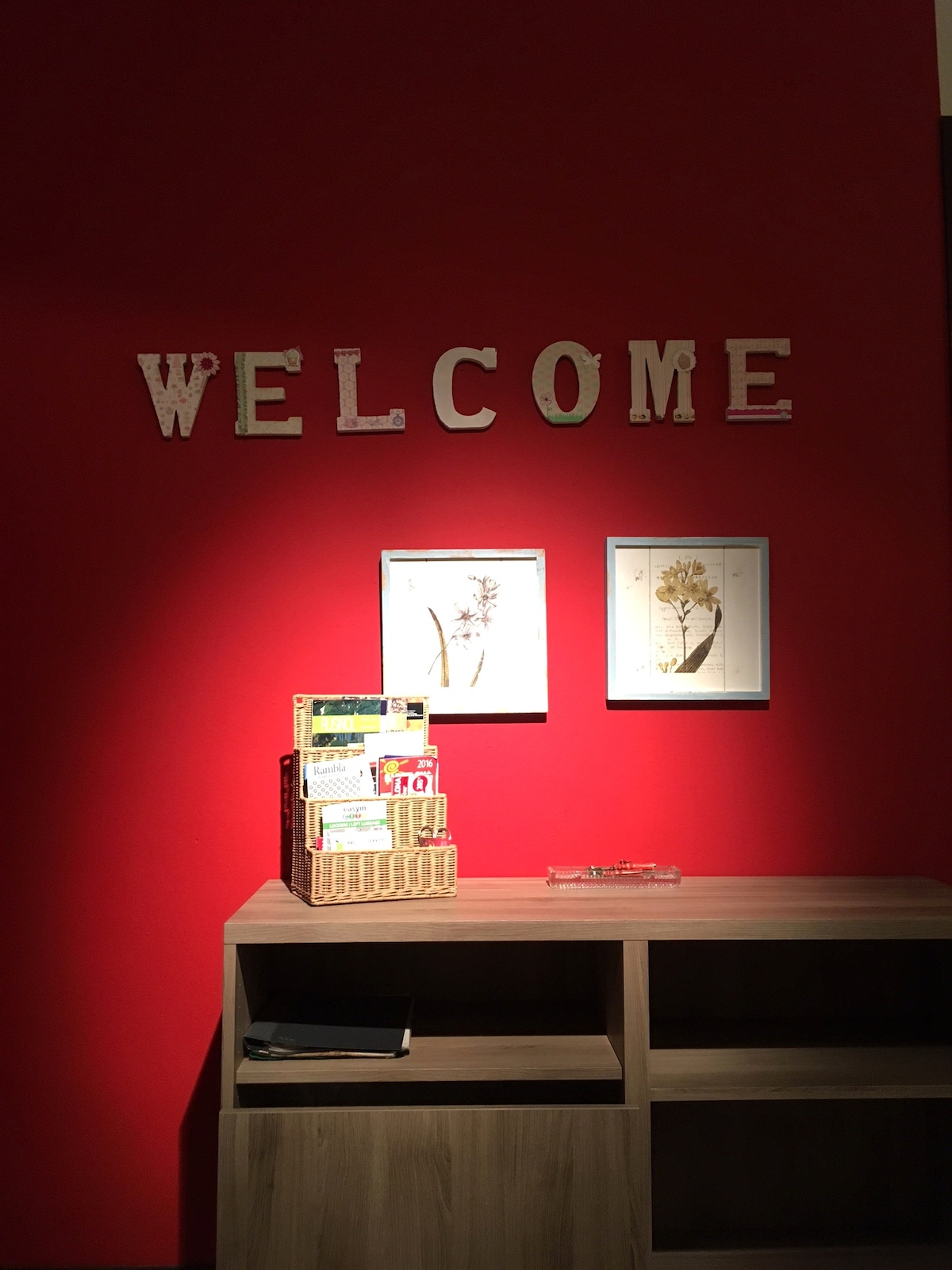 A bright red wall displays the word 'WELCOME' in white letters. Below, two framed botanical prints are positioned symmetrically. A wooden shelving unit hosts a woven basket filled with various materials, complemented by a clear tray, creating an inviting entryway.