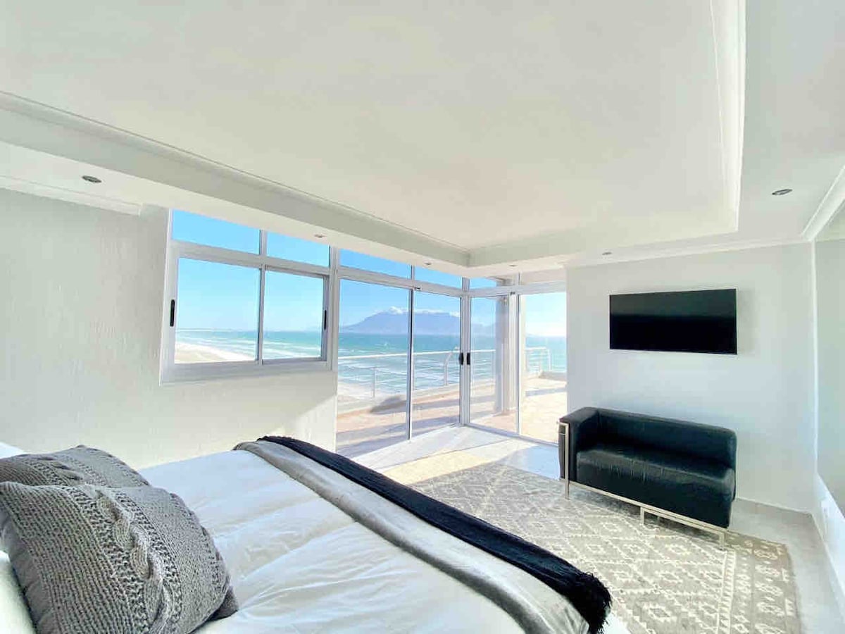 A serene bedroom features a spacious bed adorned with soft linens. Large windows provide panoramic views of the ocean and Table Mountain, filling the room with natural light. A sleek black couch is positioned near the window, and a flat-screen TV is mounted on the wall.