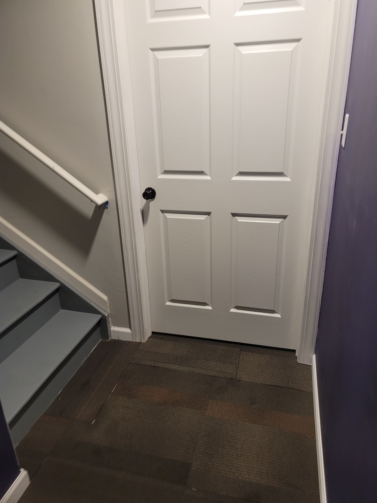 A staircase leads to a closed white door, framed by light-colored walls. The stairs are painted in a soft blue hue, while the floor features a mix of dark and light tiles. A handrail provides support alongside the steps.