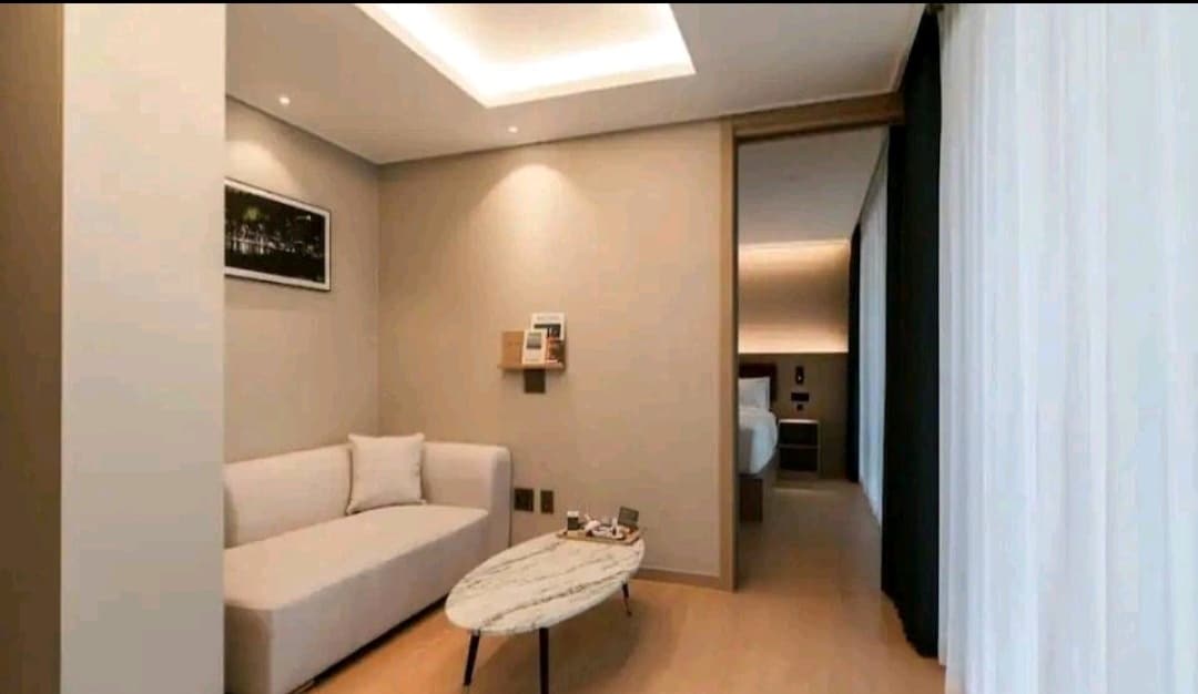 A modern living space is presented, featuring a light-colored sofa and a marble coffee table. Soft lighting highlights the neutral walls, while large windows allow natural light to fill the room. The layout connects to a bedroom area, offering a spacious and clean atmosphere.