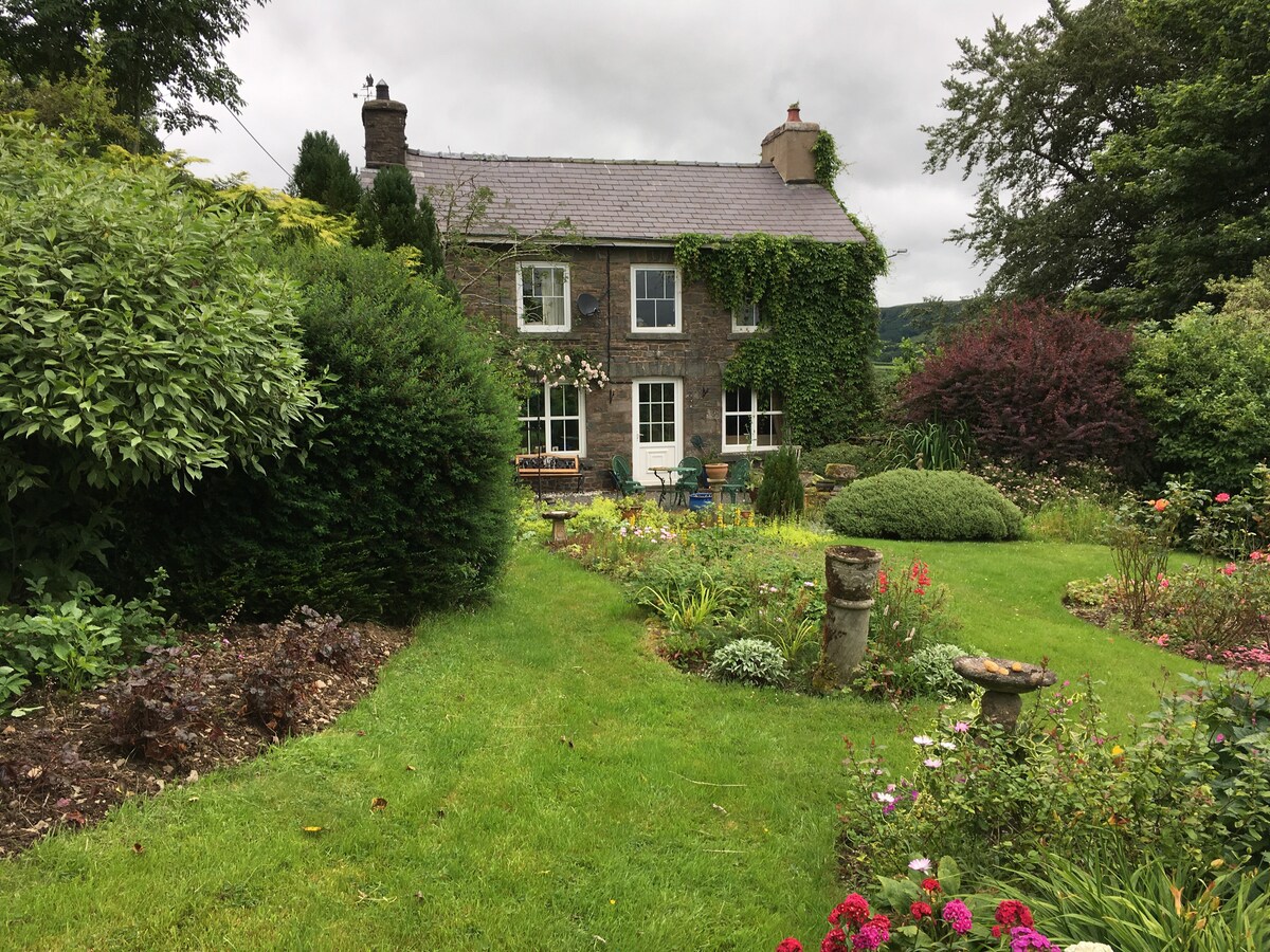 A charming stone cottage is surrounded by a lush, mature garden filled with various plants and flowers. The exterior features multiple windows, which allow natural light to enter, while the garden paths and seating areas invite relaxation amidst the greenery.