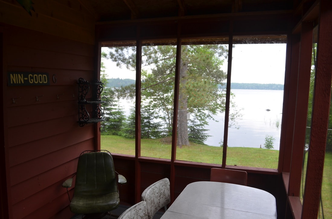 A screened-in porch offers a view of Farm Lake, framed by trees and greenery. A rustic table and two chairs sit beneath a wooden ceiling, while a coat rack is mounted on the wall, enhancing the inviting atmosphere.