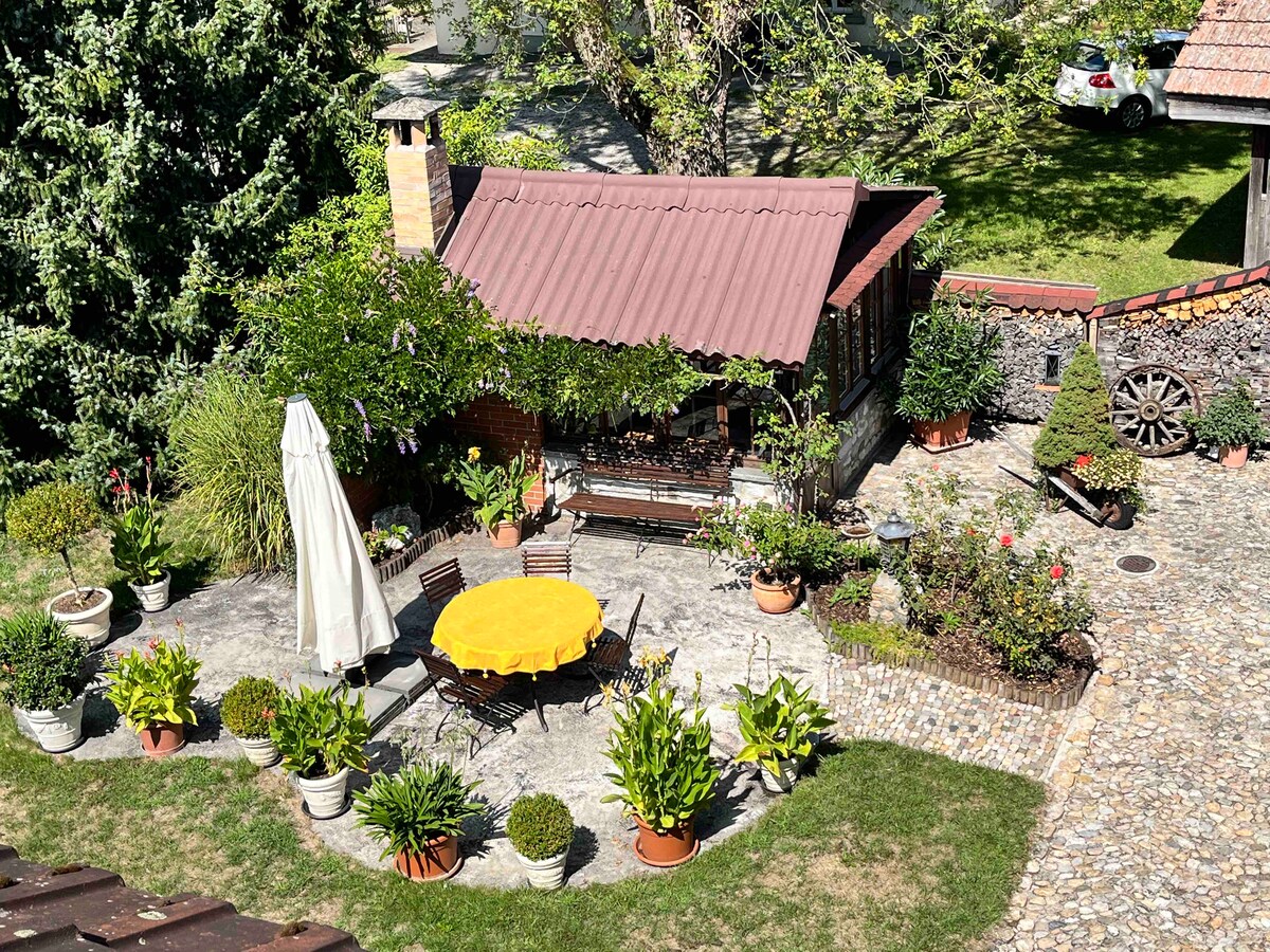 A serene outdoor area features a round table with a bright yellow tablecloth, surrounded by wooden chairs. Lush greenery, potted plants, and flowers create a vibrant setting. A shaded gazebo offers additional seating, while cobblestone paths and a rustic stone wall enhance the inviting atmosphere.