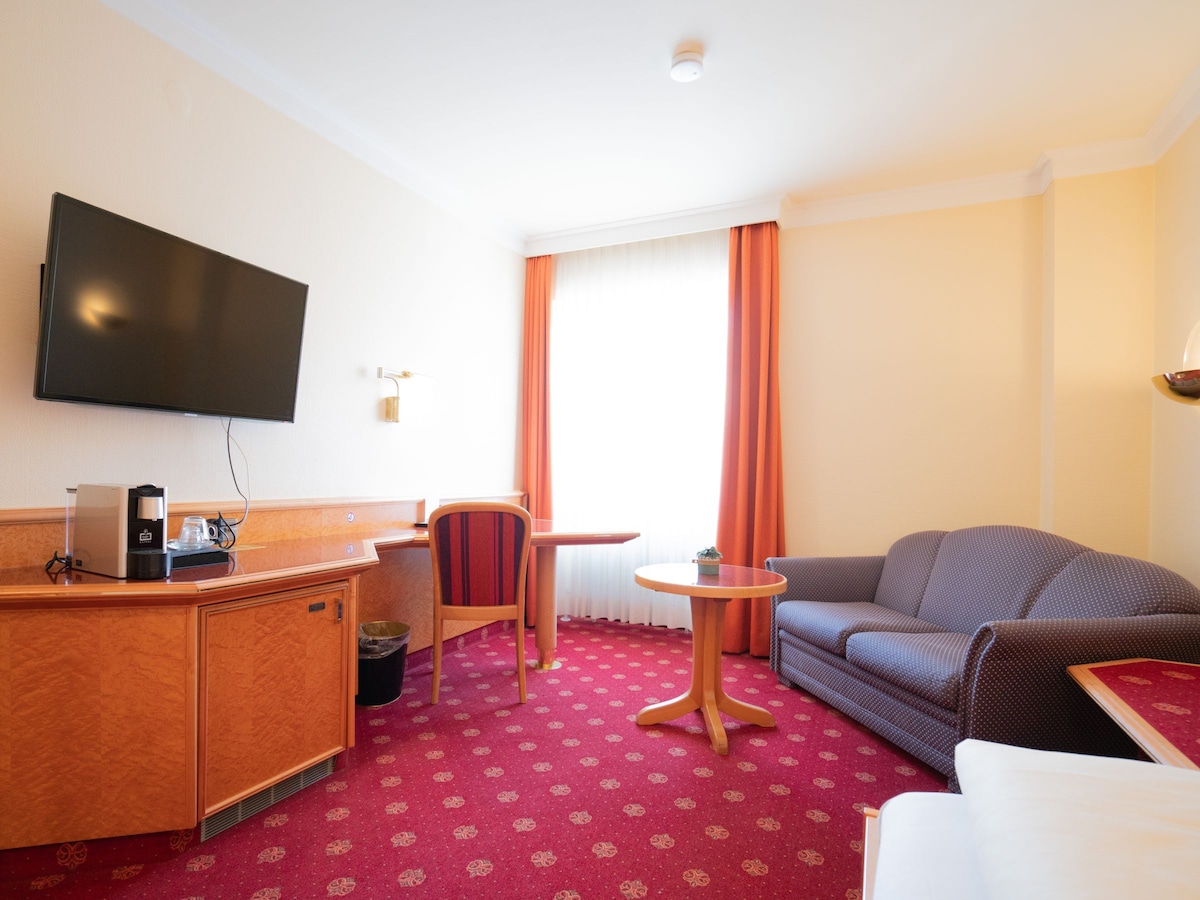 A cozy room features a comfortable seating area with a dark sofa and a round coffee table. A desk is positioned alongside a flat-screen TV mounted on the wall. Natural light streams through a large window, adorned with light curtains, enhancing the warm decor.