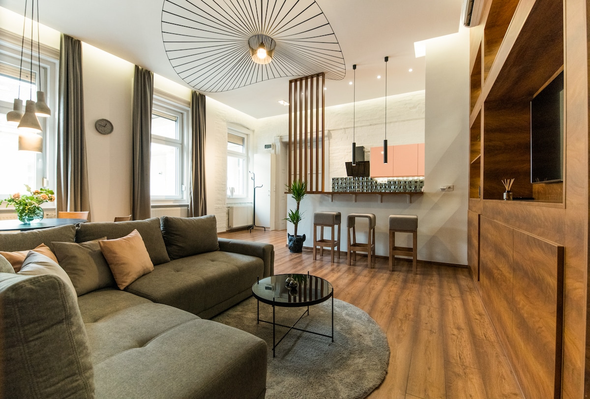 A modern living area features a comfortable sectional sofa complemented by decorative pillows. A round black coffee table rests on a plush area rug, while a sleek bar area with high stools is visible in the background. Large windows allow natural light, enhancing the open layout.