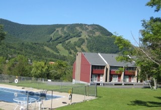 A view of the condo complex is captured against a backdrop of a lush green mountain. The outdoor pool is visible in the foreground, surrounded by a fenced area, with a picnic area and the well-maintained lawn enhancing the tranquil setting.