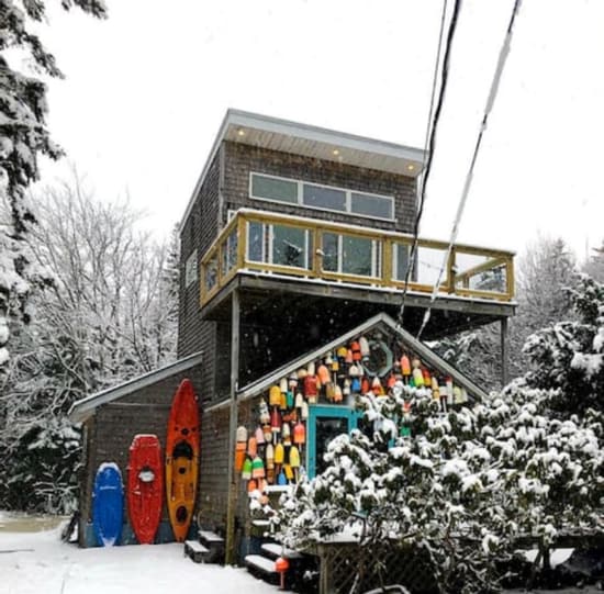 A two-story cottage is surrounded by snow, featuring a colorful display of buoys on one side. A deck with glass railing overlooks the winter landscape from the upper level, while the lower floor has a vibrant turquoise door amidst greenery.