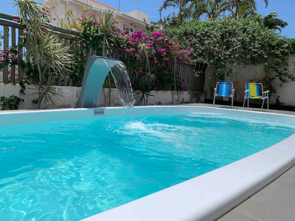 A refreshing swimming pool is framed by vibrant greenery and blooming plants. Water cascades gently from a modern feature into the clear water, inviting relaxation. Two colorful lounge chairs are positioned nearby, creating a serene outdoor space for guests.