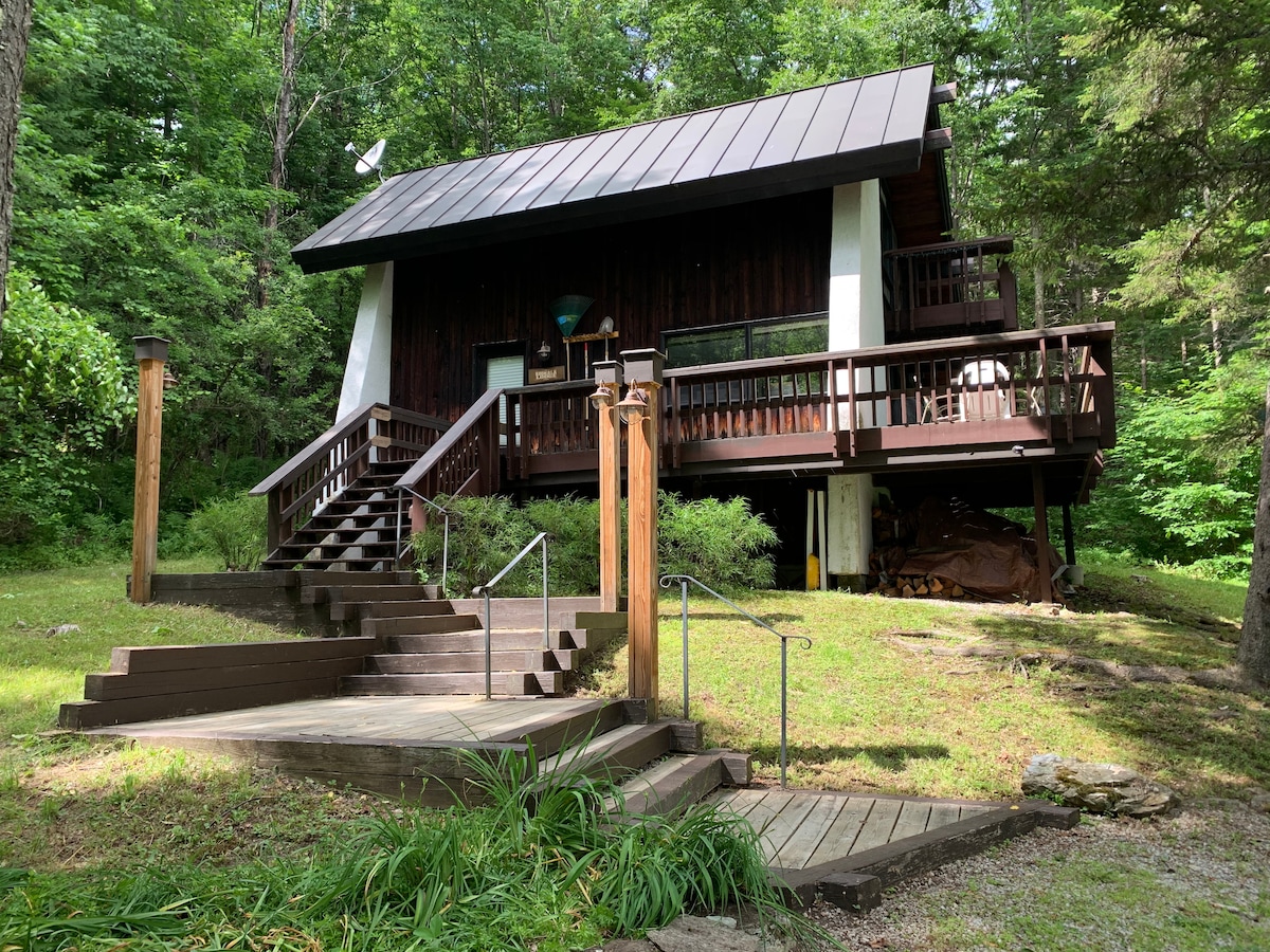 A private house is nestled in a wooded area, featuring a dark wood exterior and a metal roof. Steps lead to a wraparound deck, and nearby pathways are accented with natural landscaping. The setting offers a tranquil atmosphere surrounded by trees.