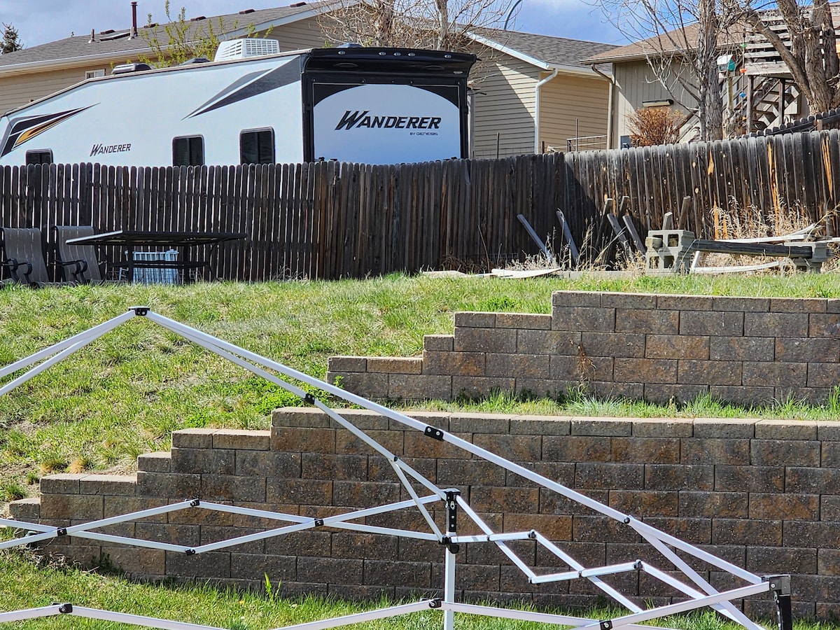 A recreational vehicle is parked behind a wooden fence, showcasing its side marked with the word 'Wanderer.' In the foreground, a partially assembled canopy frame is visible on a grassy area with tiered stone steps leading upward.