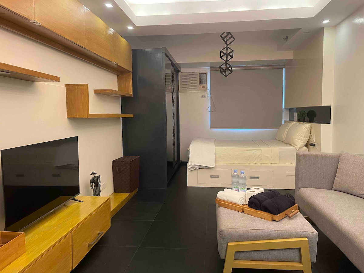 A compact living space features a cozy bed positioned against the wall, complemented by a television and a light wooden shelf. A small seating area includes a grey sofa with a wooden tray. Natural light filters through a window covered by a roller blind.