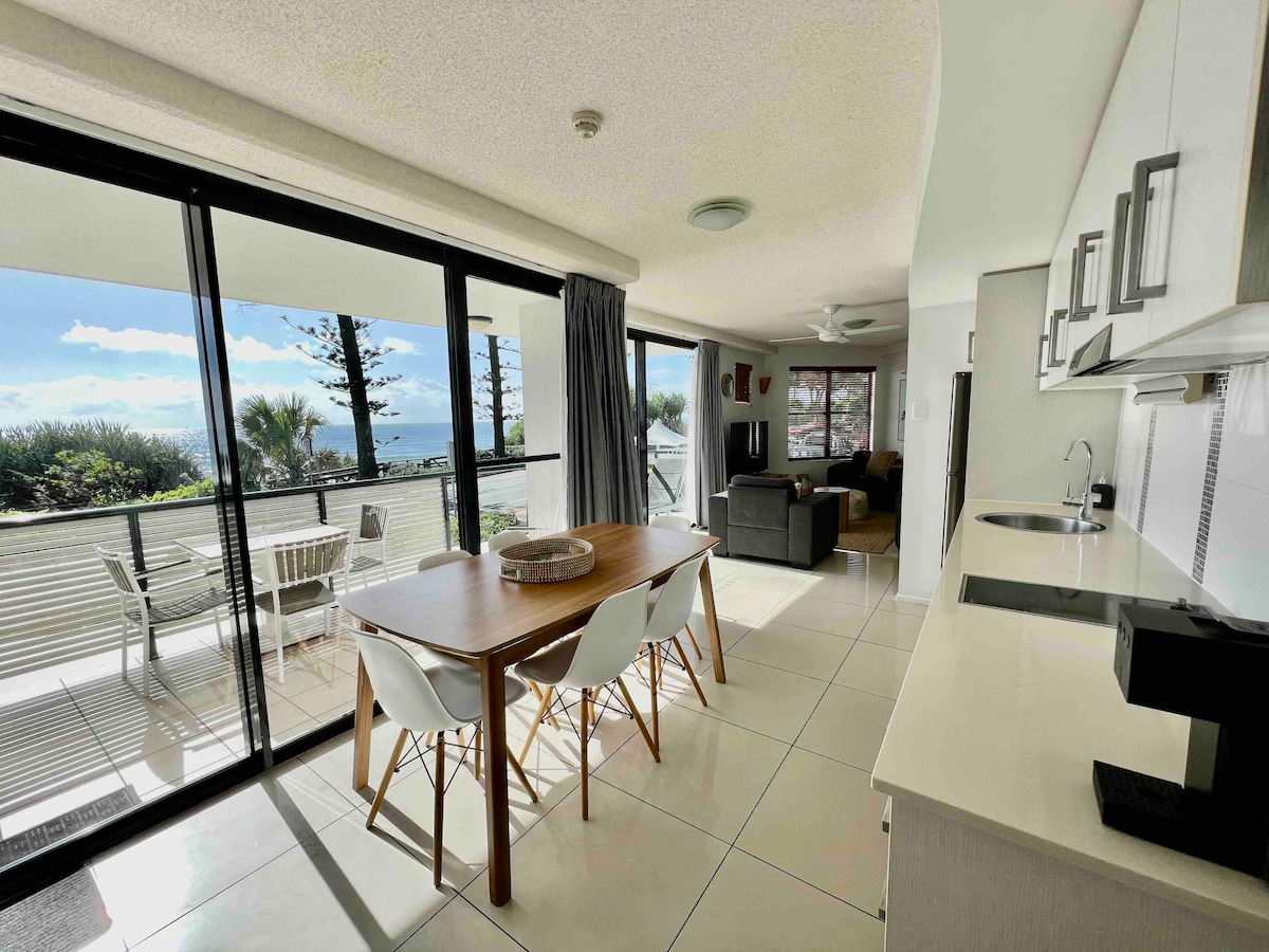 A spacious living area is showcased with large glass doors allowing natural light and offering views of the ocean. The dining table is paired with modern chairs, while a comfortable sofa sits in the background. A kitchenette is visible, featuring sleek cabinetry and appliances.