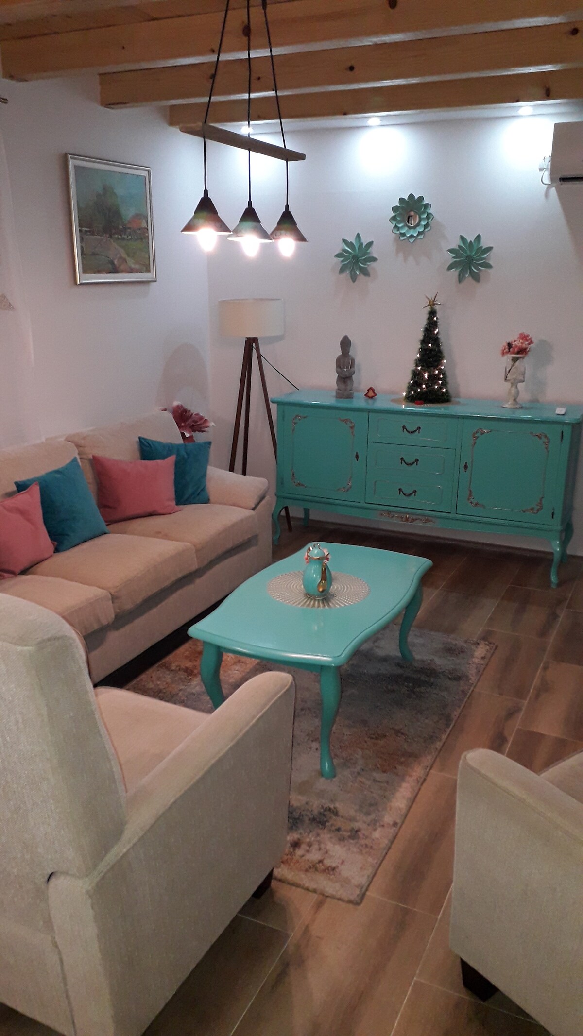 A cozy living area features a light-colored sofa paired with accent chairs. A turquoise coffee table is centered on a soft area rug, while decorative elements include a small decorative piece and unique wall art. Soft lighting is provided by hanging light fixtures.