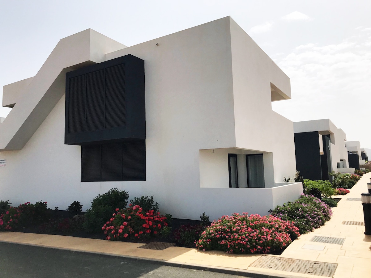 A modern white building with geometric lines is set within a landscaped area filled with vibrant flowering plants. Large windows provide natural light to the interior. The surrounding pathway and garden enhance the minimalist aesthetic of the urbanization.