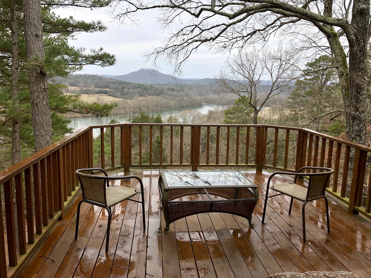 A wooden deck overlooks a scenic view of rolling hills and a river. Two chairs are positioned around a glass-topped table, while the wet surface of the deck reflects the surrounding natural landscape, featuring trees and distant mountains.