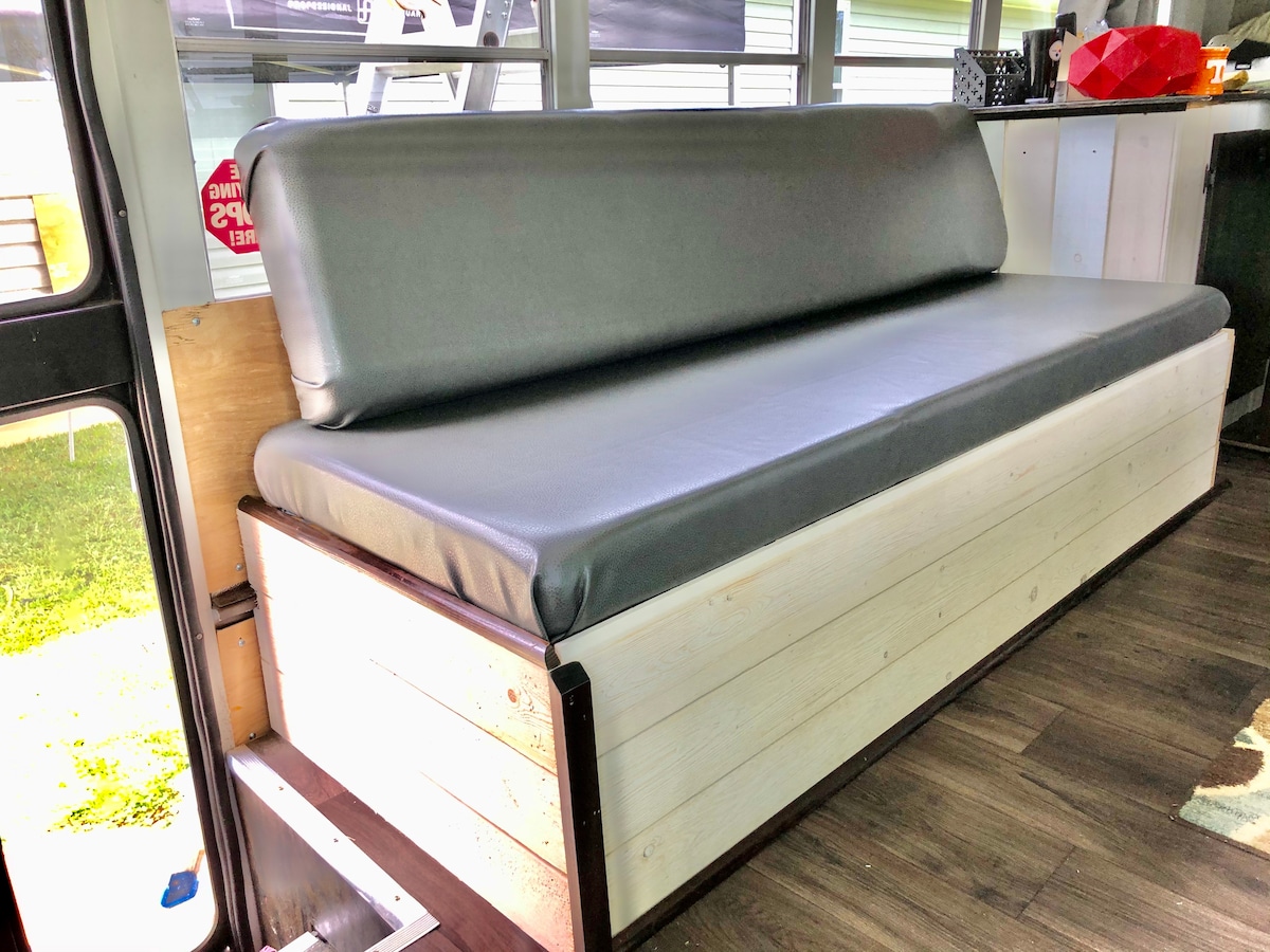 A comfortable couch is positioned along the interior wall of the converted camper bus. The seating features a smooth, dark grey cushion atop a wooden base, providing a cozy spot for relaxation. Natural light streams through a nearby window, illuminating the space.