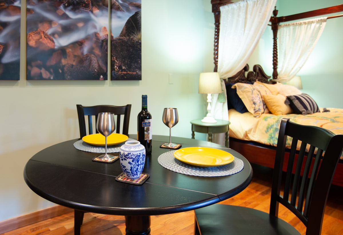 A cozy dining area is set with a black table and matching chairs, featuring two wine glasses and yellow dinner plates. In the background, a carved mahogany four-poster queen bed is seen, adorned with decorative pillows and softly illuminated by bedside lamps.