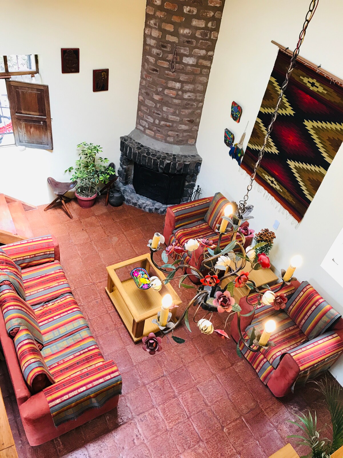 A spacious living area is visible, featuring two vibrant sofas adorned with colorful stripes. A rustic stone fireplace serves as a focal point, while a unique chandelier hangs above a wooden coffee table. Cultural artwork adds warmth to the inviting space.