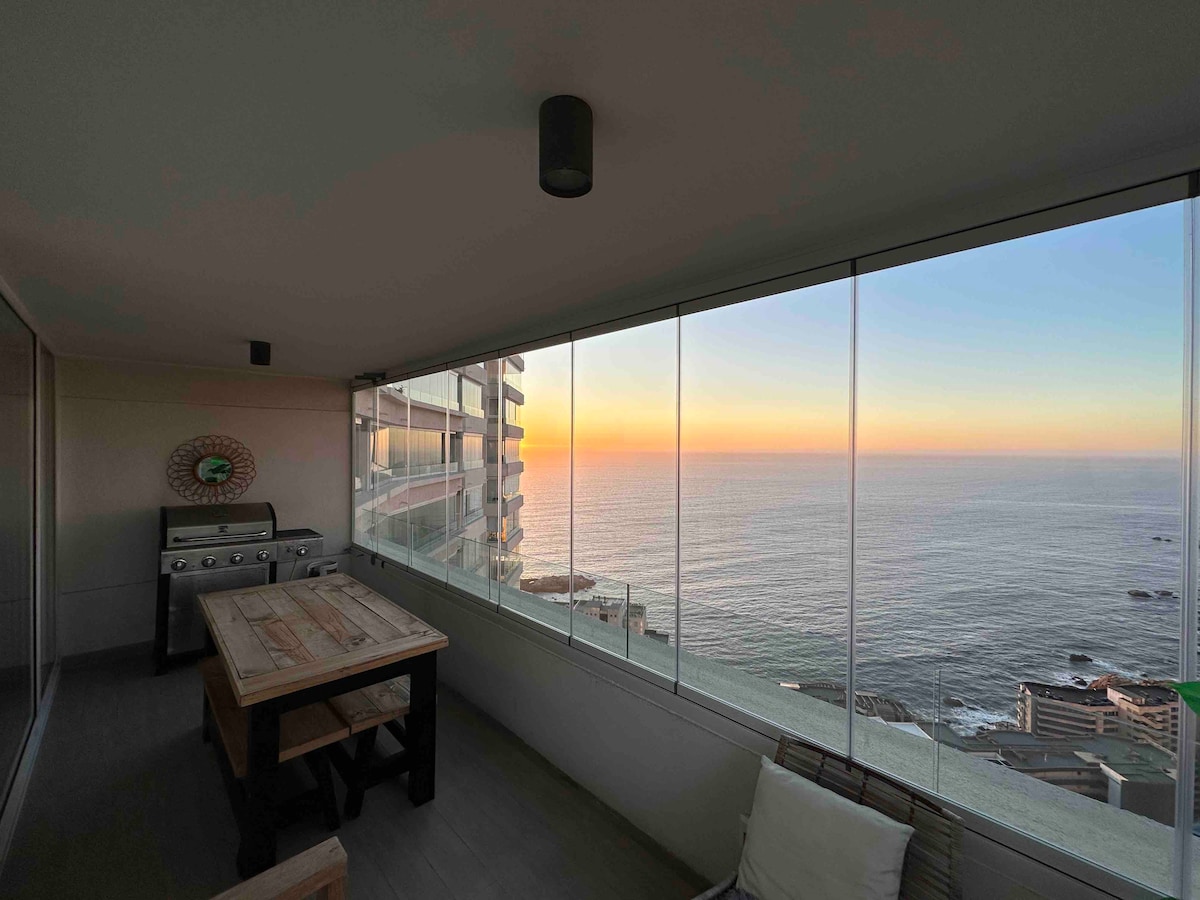 A sunlit terrace offers a panoramic view of the ocean, with large glass panels providing an unobstructed sight of the horizon. A wooden table is positioned under a soft overhead light, while a grill is located nearby, inviting al fresco dining experience during sunset.