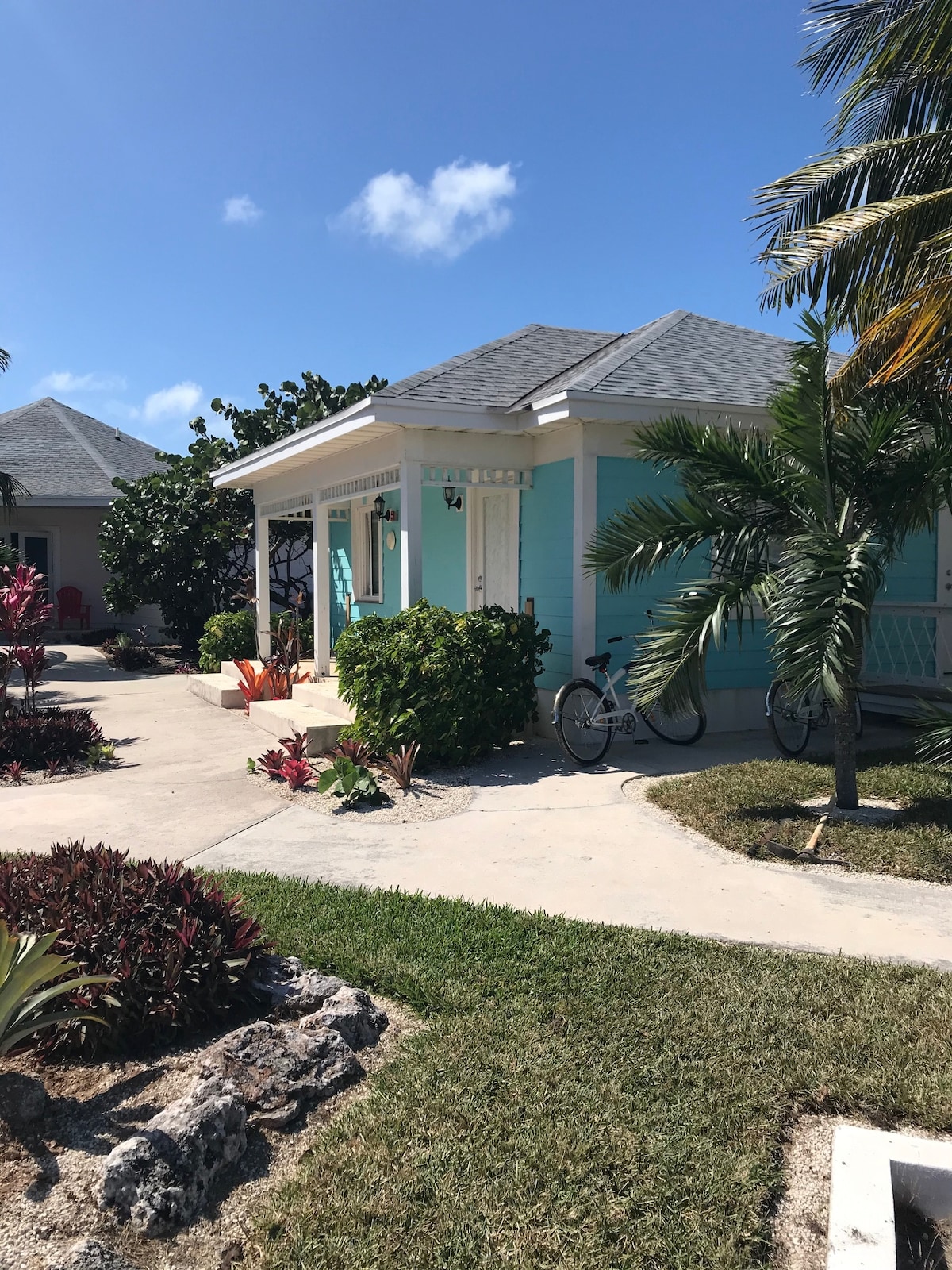 A charming exterior view of the accommodation features a light blue facade surrounded by lush green landscaping. A bicycle is parked nearby on the pathway, and tropical plants add a vibrant touch to the setting. A clear blue sky is visible overhead.