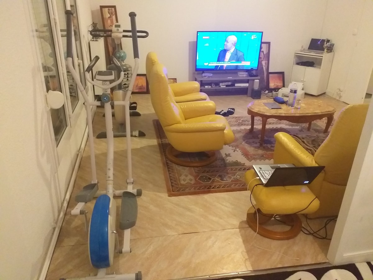 A living room area features two yellow armchairs facing a television displaying a news program. A small wooden coffee table is positioned in the center, surrounded by a plush area rug. An exercise bike is placed near a window, and a laptop rests on a side table.