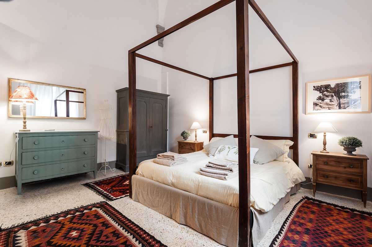 A spacious bedroom features a four-poster bed with soft linens and two bedside tables. A vintage dresser and a wardrobe complement the room, while decorative rugs add warmth to the flooring. Gentle lighting from table lamps enhances the serene ambiance.