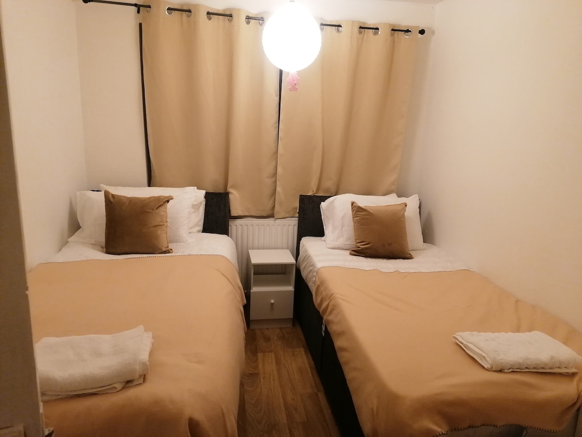 A well-lit bedroom features two neatly made single beds, each dressed in light beige covers. Soft brown decorative pillows are placed on the beds. Light curtains frame the window, and a simple nightstand is located between the beds, providing a cozy and functional sleeping space.