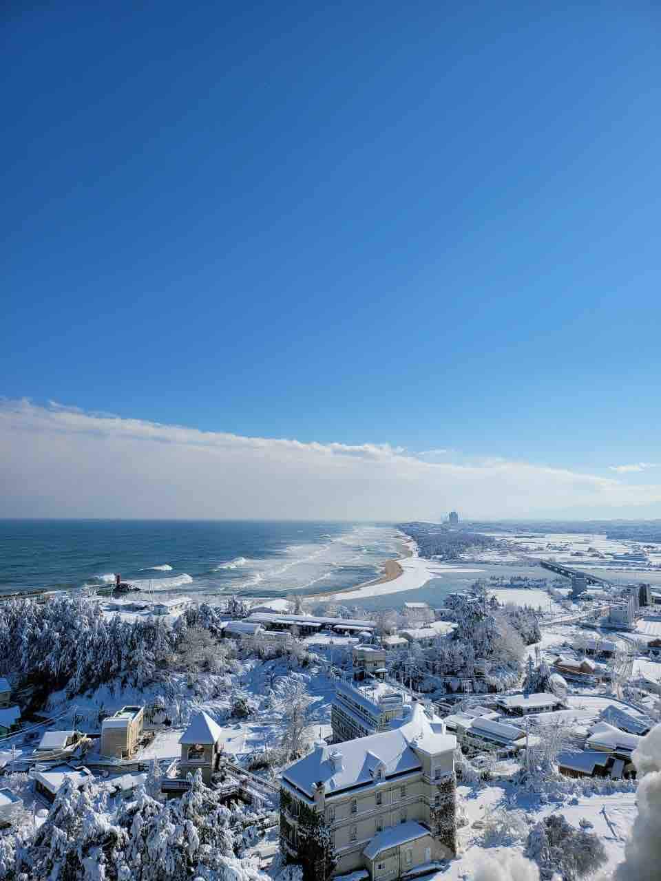 A panoramic view showcases a snow-covered landscape meeting the ocean. Pristine white buildings are contrasted against the blue water and sky. Gentle waves roll onto the sandy beach, while the distant shoreline is marked by a blend of natural and urban elements.