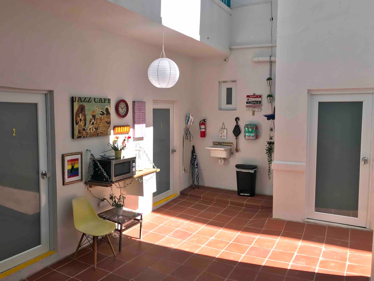A sunny courtyard showcases modern doorways leading to guest rooms. Decor includes a wall clock, framed art, and a small table with a microwave. A light fixture hangs overhead, and a trash bin is positioned near a shelf adorned with plants and local artwork.