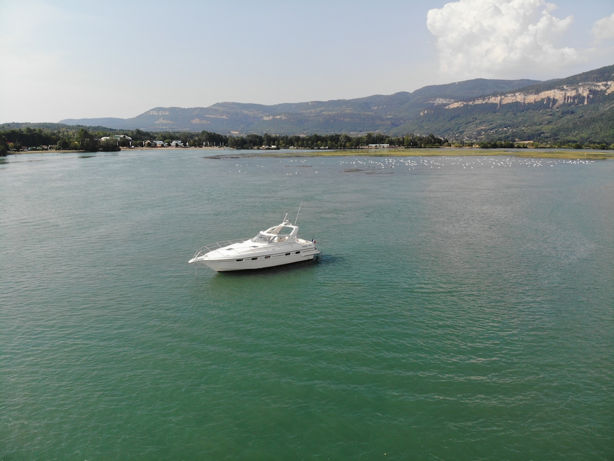 A sleek white boat is anchored in the calm waters, surrounded by verdant hills and a clear blue sky. The smooth surface of the water reflects the landscape, creating a serene atmosphere. Fluffy clouds drift lazily overhead, enhancing the tranquil setting.