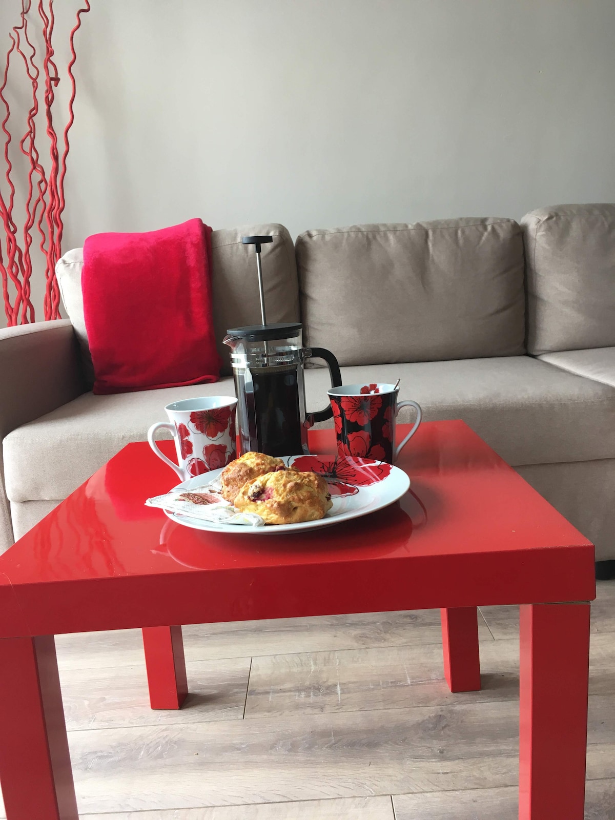 A bright red table hosts a plate of freshly baked pastries alongside two patterned mugs and a French press coffee maker. A comfortable beige sofa with a red accent pillow serves as a backdrop, creating a cozy dining setup for enjoying breakfast.