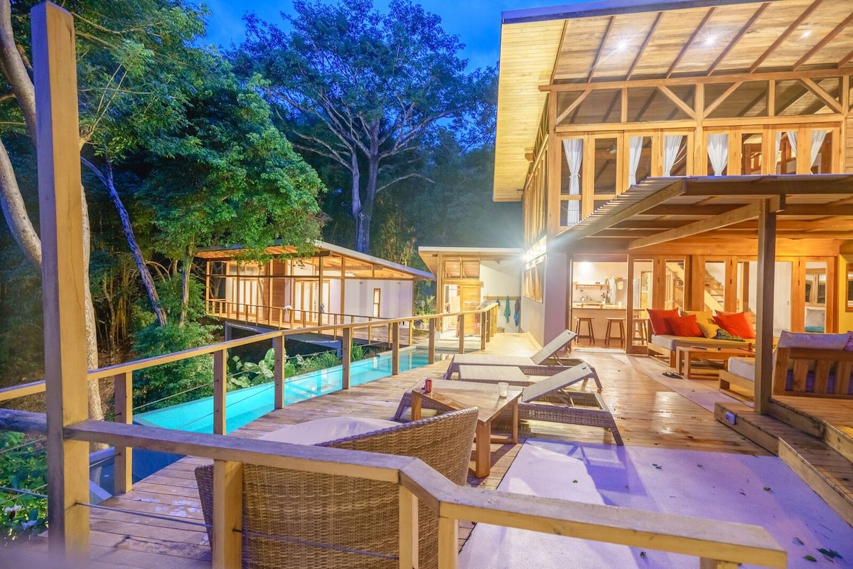 A wooden terrace is illuminated in soft evening light, showcasing multiple lounge chairs and a modern exterior. The background reveals lush greenery and a portion of an infinity pool that overlooks the surrounding jungle, enhancing the tranquil outdoor space.
