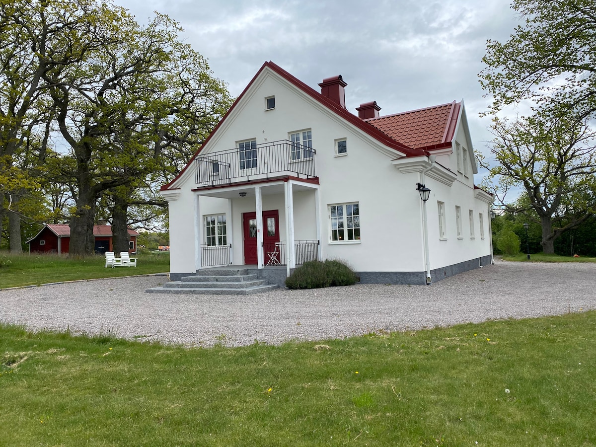 The exterior of the white, two-story house features a red roof and red accents around the doors and windows. A welcoming entrance is visible, with steps leading to a small balcony on the upper level. Surrounding greenery enhances the property’s spacious lawn.