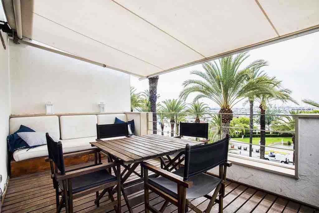 A spacious terrace is equipped with a wooden dining table surrounded by four chairs. A cozy seating area features a cushioned bench, offering views of palm trees and the surrounding park area. The space is shaded by a retractable awning, suitable for outdoor relaxation.