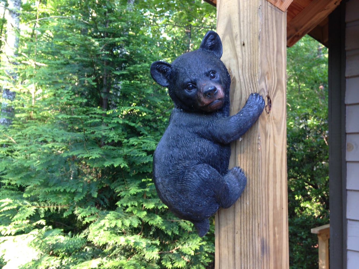 A decorative black bear sculpture is positioned on a wooden post, appearing to climb. Green foliage and trees in the background create a natural setting that enhances the cabin ambiance.