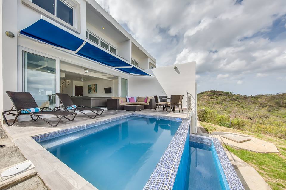 A private pool is situated outside, reflecting the surrounding landscape. Lounge chairs are positioned nearby under a blue awning. The exterior features a modern design with large glass doors that provide a view of the interior space, enhancing the indoor-outdoor connection.