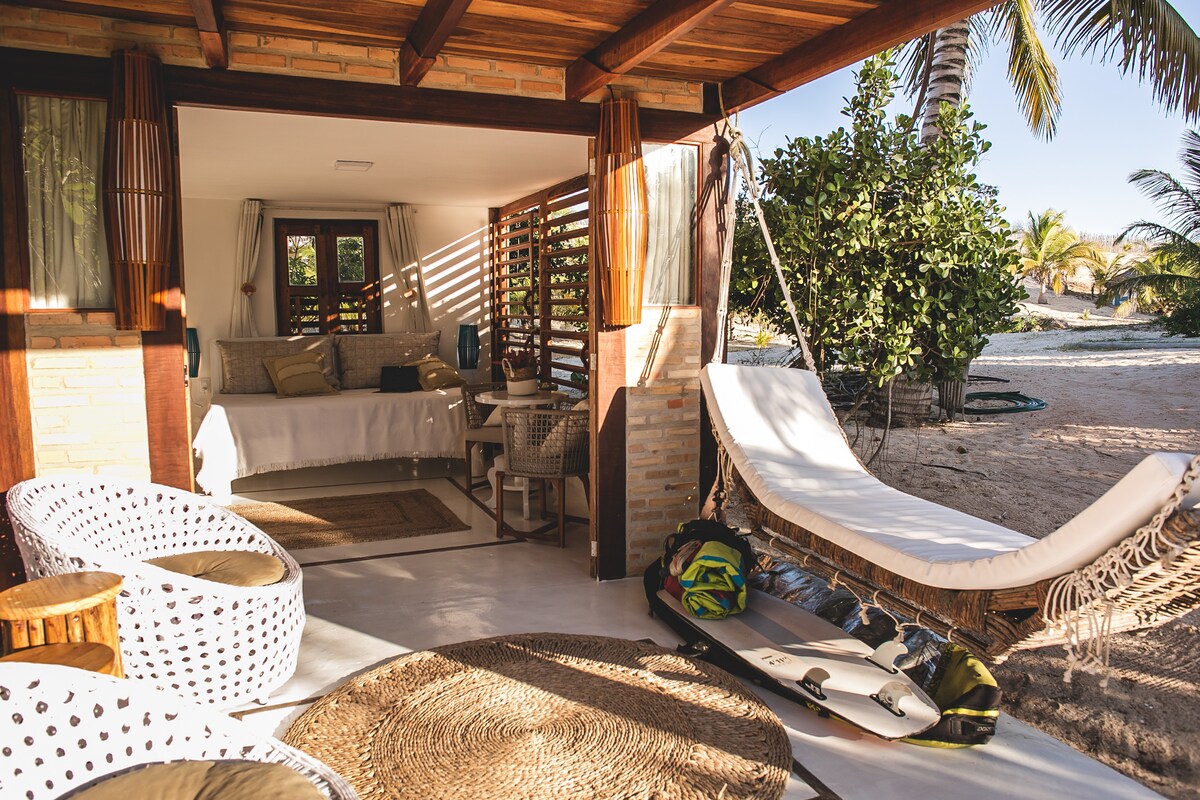 An outdoor seating area features two woven chairs and a hanging hammock, inviting relaxation. A sliding door reveals a bright indoor space with a comfortable bed and work desk. Natural light filters through slatted wood accents, creating a connection with the surrounding greenery.